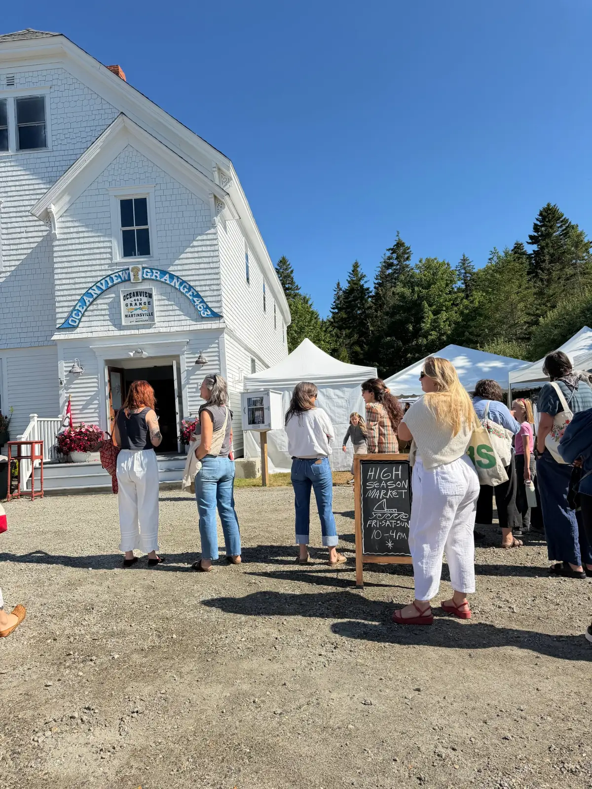 People queuing at an outdoor market beside a white clapboard hall, with a chalkboard sign