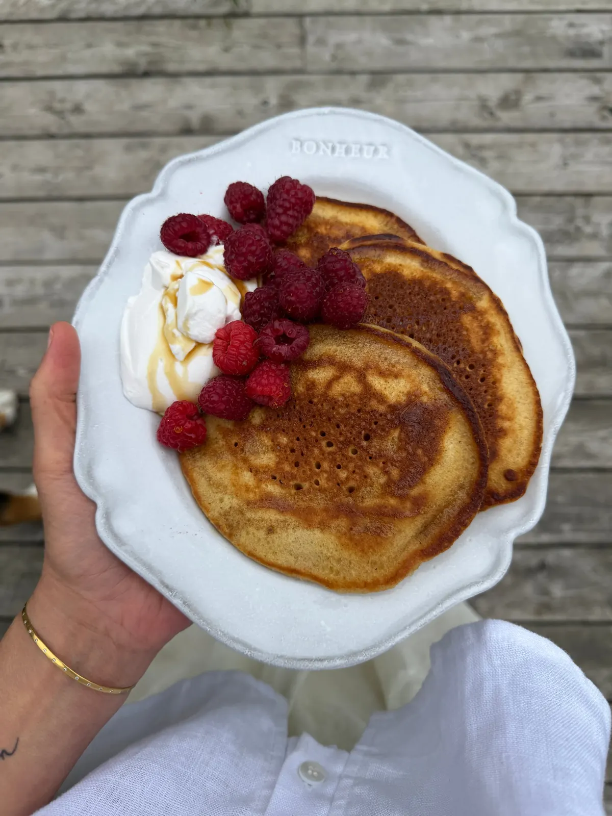 A hand holding a white plate of pancakes with raspberries and cream over a wooden deck